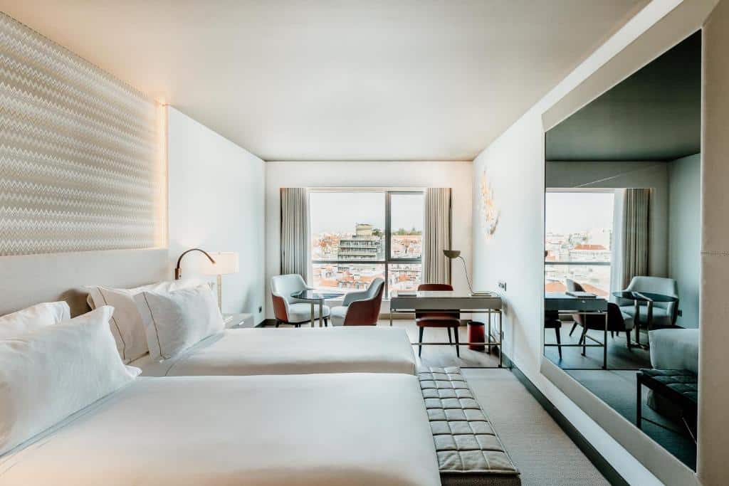 Quarto do EPIC SANA Marquês Hotel com duas camas de solteiro, uma varanda com cortinas com vista para a cidade, perto da janela há uma pequena mesinha com suas poltronas e uma mesa de escritório, além de um amplo espelho de frente para as camas, para representar hotéis perto da Praça Marquês de Pombal em Lisboa