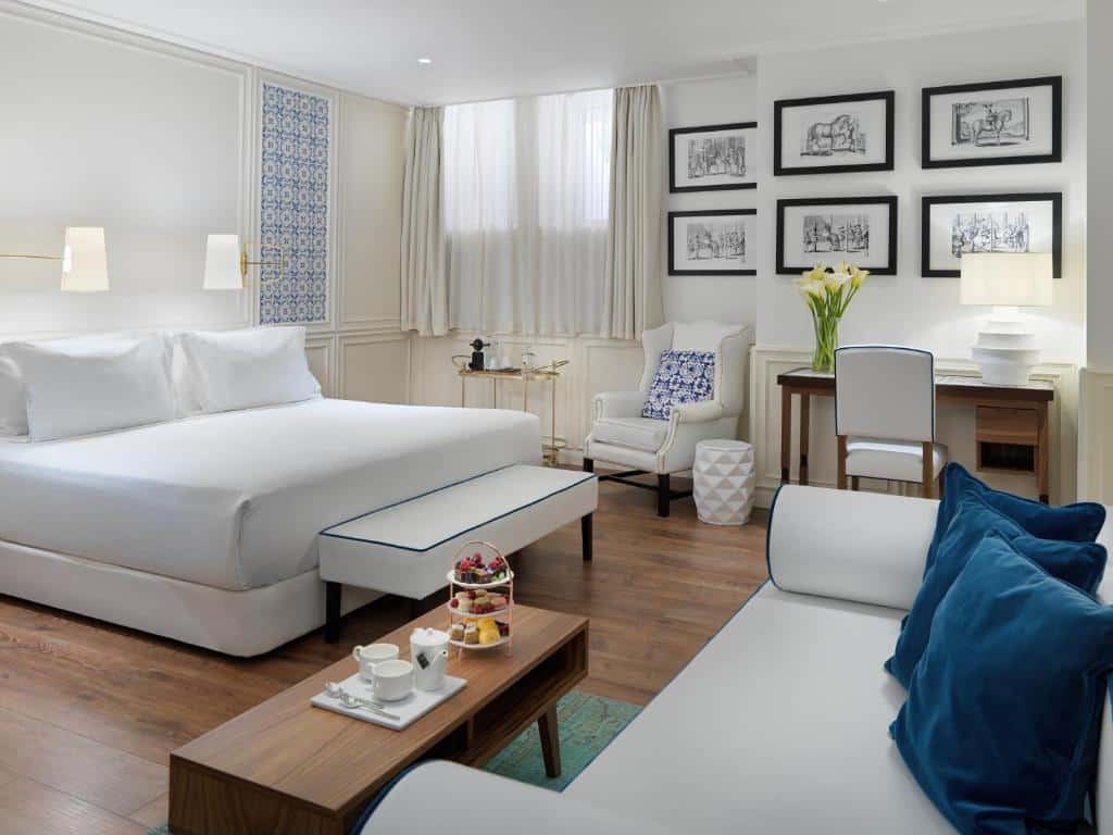 Quarto amplo do H10 Duque de Loule com uma cama de casal, janela com cortinas, quadros decorando o ambiente, uma pequena mesa de madeira com um abajur e um vaso de flor em cima, há também um sofá com almofadas e uma poltrona no ambiente
