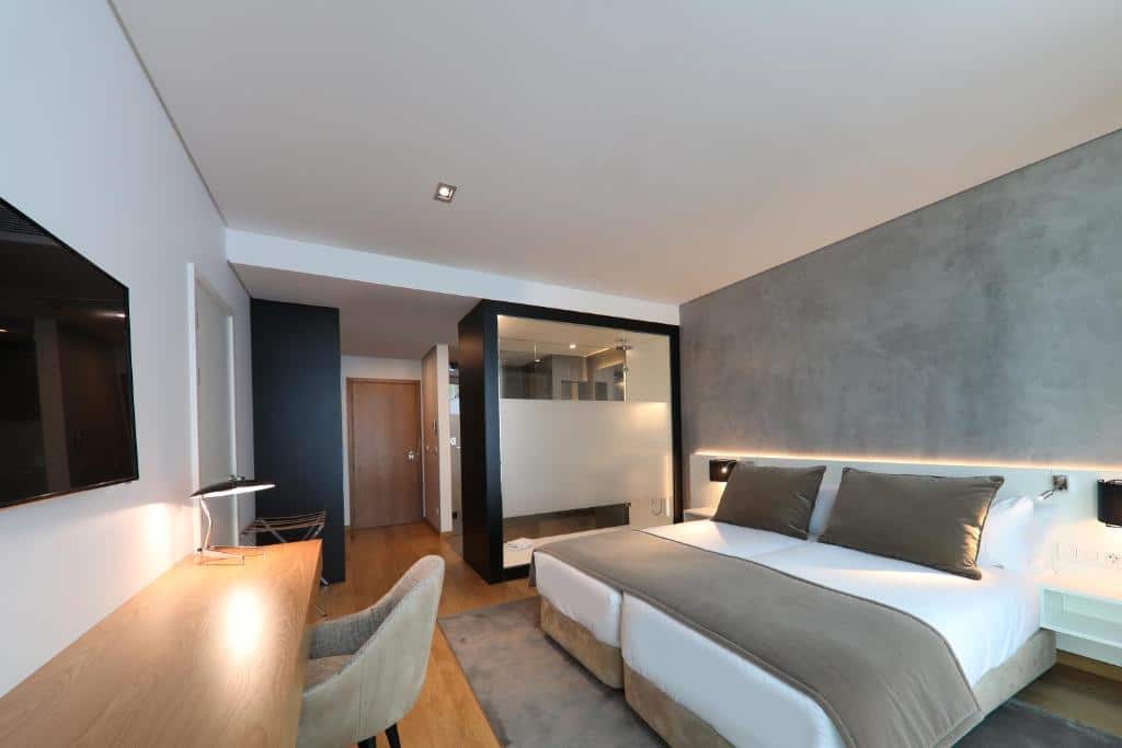 Quarto do Iberostar Selection Lisboa com uma cama de casal, uma janela que vai do teto ao chão com vista para a cidade, ao lado dessa janela há uma poltrona estofada e do lado direito está uma cama de casal com mesinhas de cabeceira e luminárias, para representar hotéis perto da Praça Marquês de Pombal em Lisboa