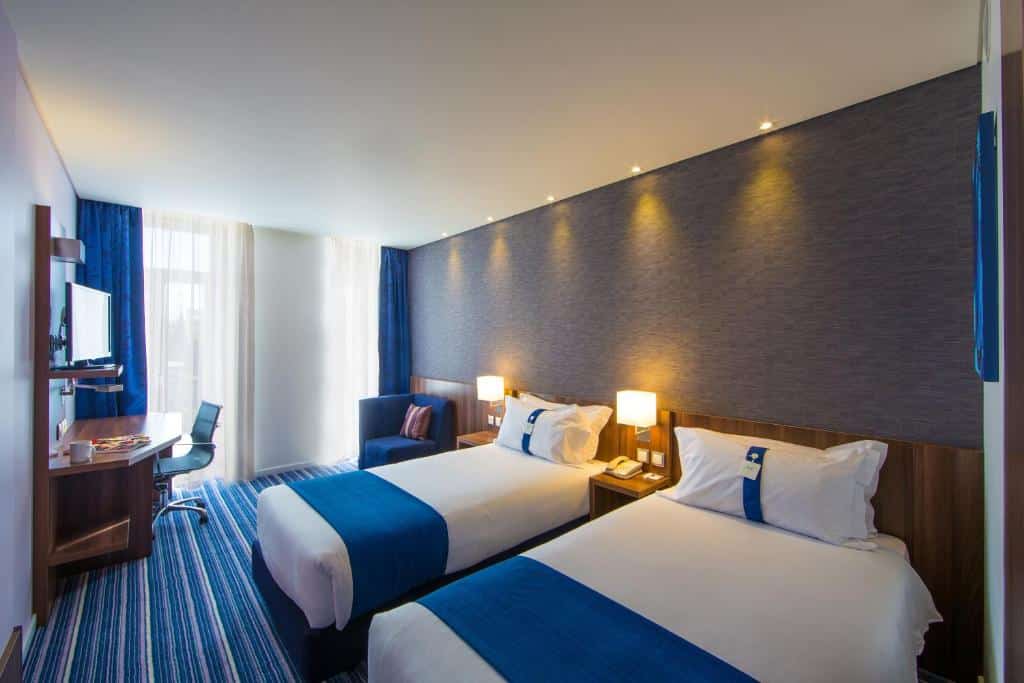 Quarto do Holiday Inn Express Lisboa - Av. Liberdade, an IHG Hotel com duas camas de solteiro, duas pequenas sacadas com cortinas, próximo delas há uma poltrona, uma mesa de escritório com uma cadeira giratória, além de uma televisão, para representar hotéis perto da Praça Marquês de Pombal em Lisboa