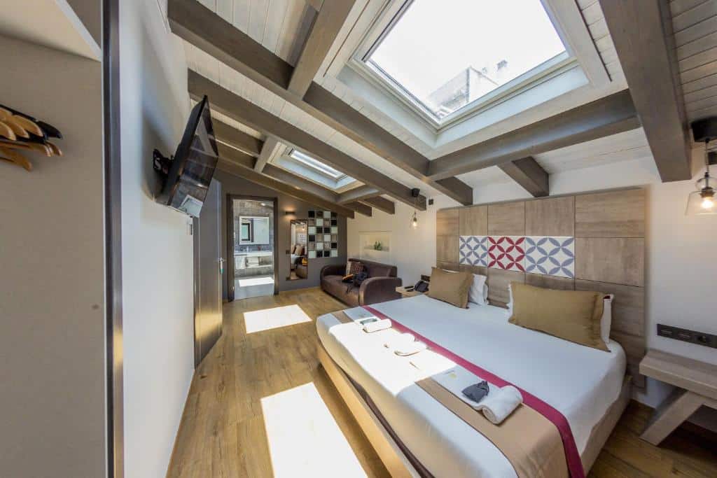 Quarto da Boutique Hostemplo Sagrada Familia, uma das recomendações de hotéis perto da Sagrada Família em Barcelona. Há janelas no teto do quarto, estando uma logo acima da cama de casal que tem mesinhas de cabeceira dos dois lados, e outra acima do sofá na área de estar ao longe. De frente para a cama há uma televisão.