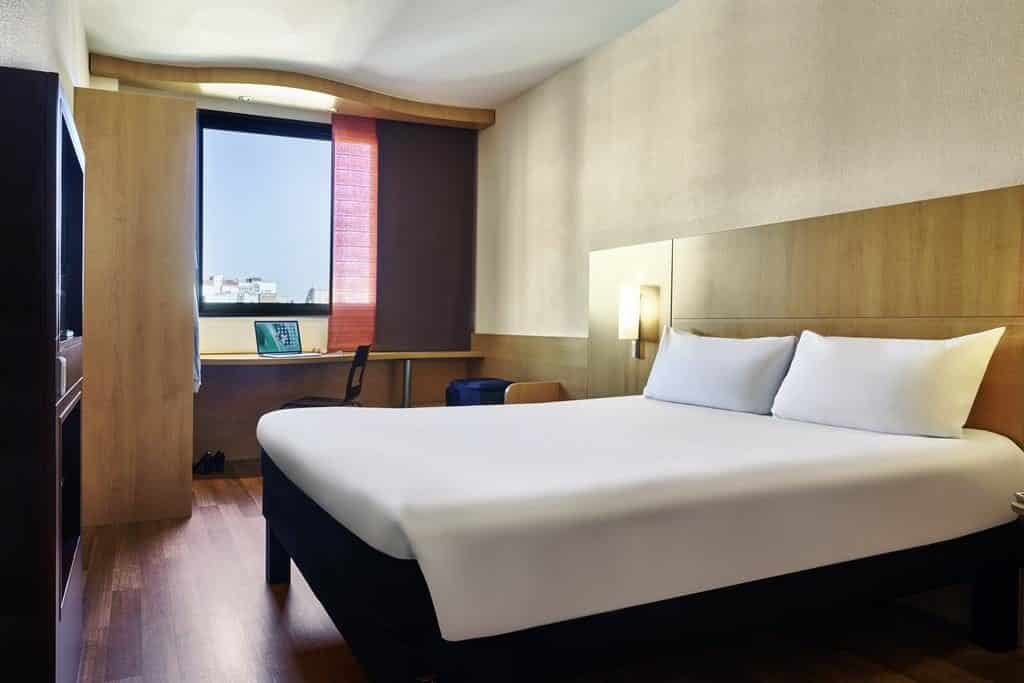 Quarto do Ibis Barcelona Centro, uma das recomendações de hotéis perto da Sagrada Família em Barcelona. A cama tem lençol e travesseiros brancos, e há luminárias dos dois lados da cabeceira de madeira.
Há uma mesa de trabalho com cadeira de frente para uma janela, e há um ármario ao lado.