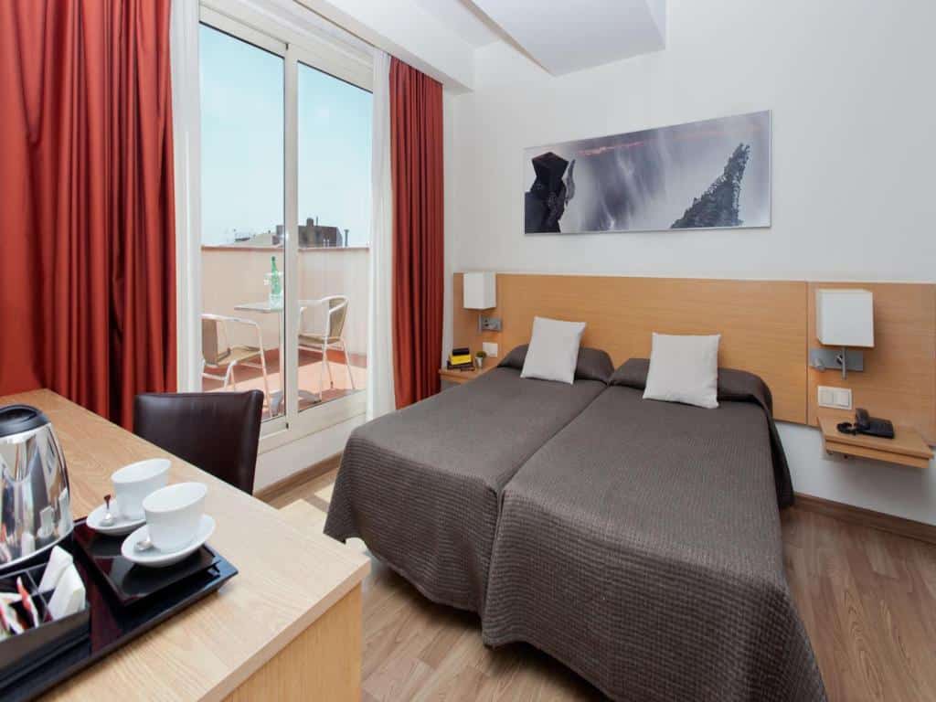 Quarto do Hotel Sagrada Família, uma das recomendações de hotéis perto da Sagrada Família em Barcelona. Duas camas de solteiro encostadas uma na outra tem mesinhas com abajur dos dois lados na cabeceira. Na parede acima há um quadro. De frente para a cama há uma mesa de trabalho com cadeira e amenidades de café. Do lado esquerdo da cama, uma porta de vidro com cortinas vermelhas tem vista para o terraço com mesa e cadeiras.