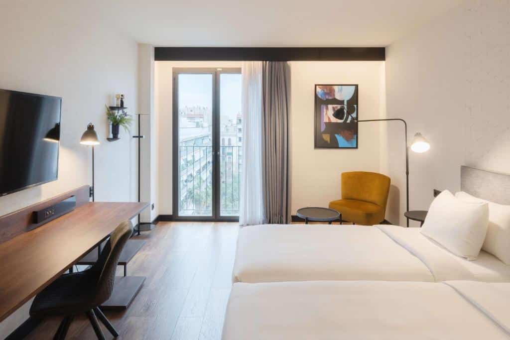 Quarto do Radisson Blu 1882 Hotel, uma das recomendações de hotéis perto da Sagrada Família em Barcelona. Duas camas de solteiro encaram uma mesa de trabalho com cadeira e televisão na parede acima. As camas têm mesinha de cabeceira com abajur dos dois lados, e na parede de frente para a foto há uma janela de vidro com vista. Ao lado, uma poltrona tem uma mesinha logo em frente.
