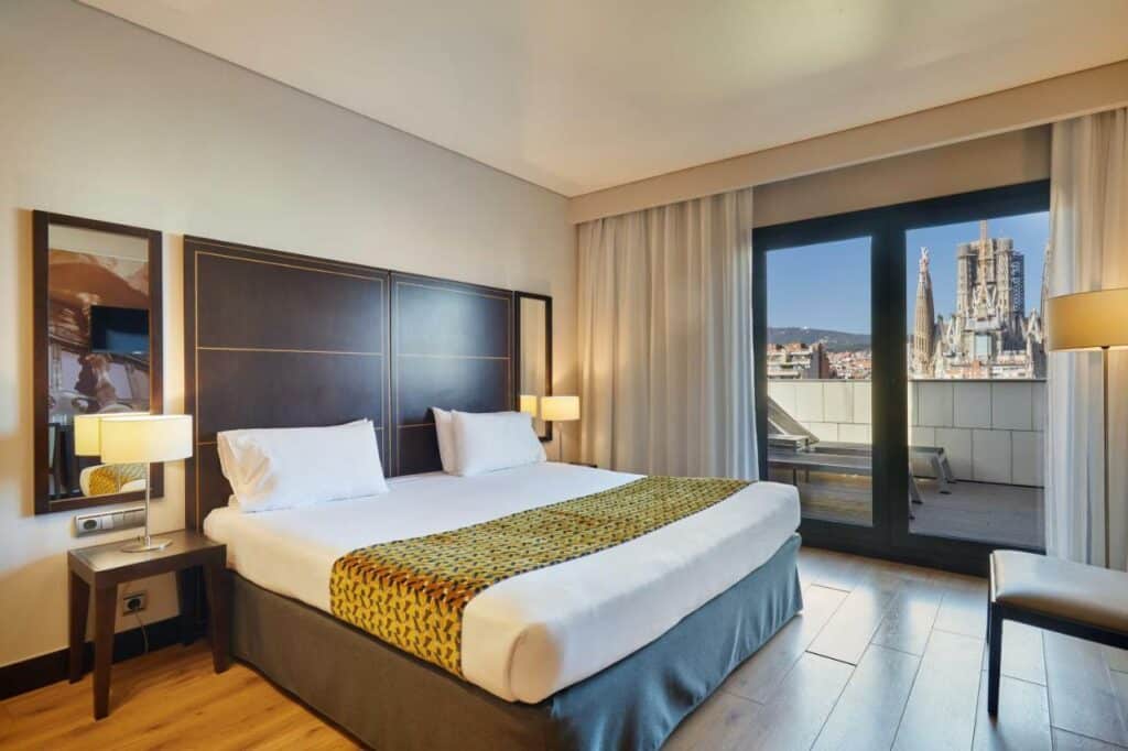 Quarto do Eurostars Monumental, uma das recomendações de hotéis perto da Sagrada Família em Barcelona. A cama de casal tem mesinhas de cabeceira com abajur e espelho dos dois lados. Ao fundo, portas de vidro duplas levam ao terraço com espreguiçadeiras e vista para a Sagrada Família.