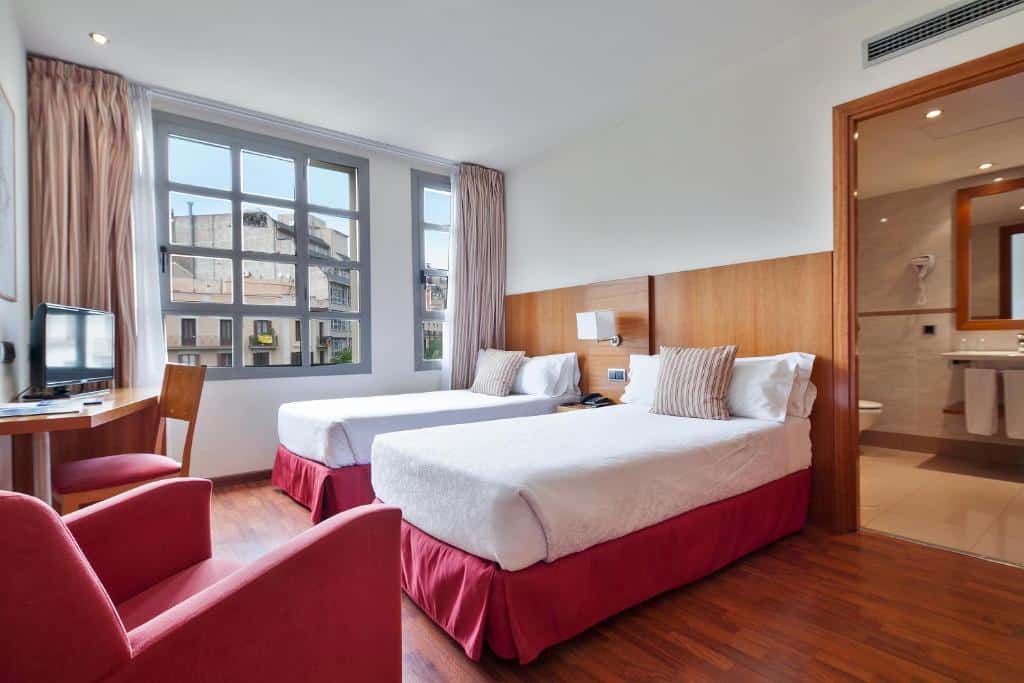 Quarto do Hotel Best Aranea, uma das recomendações de hotéis perto da Sagrada Família em Barcelona. Duas camas de solteiro tem uma mesinha de cabeceira com luminária entre si. Em frente, uma mesa com cadeira e televisão está encostada na parede com uma poltrona vermelha do outro lado. Na parede do canto, uma janela com cortinas tem vista para a rua. É possível ver a porta do banheiro no canto do quarto.