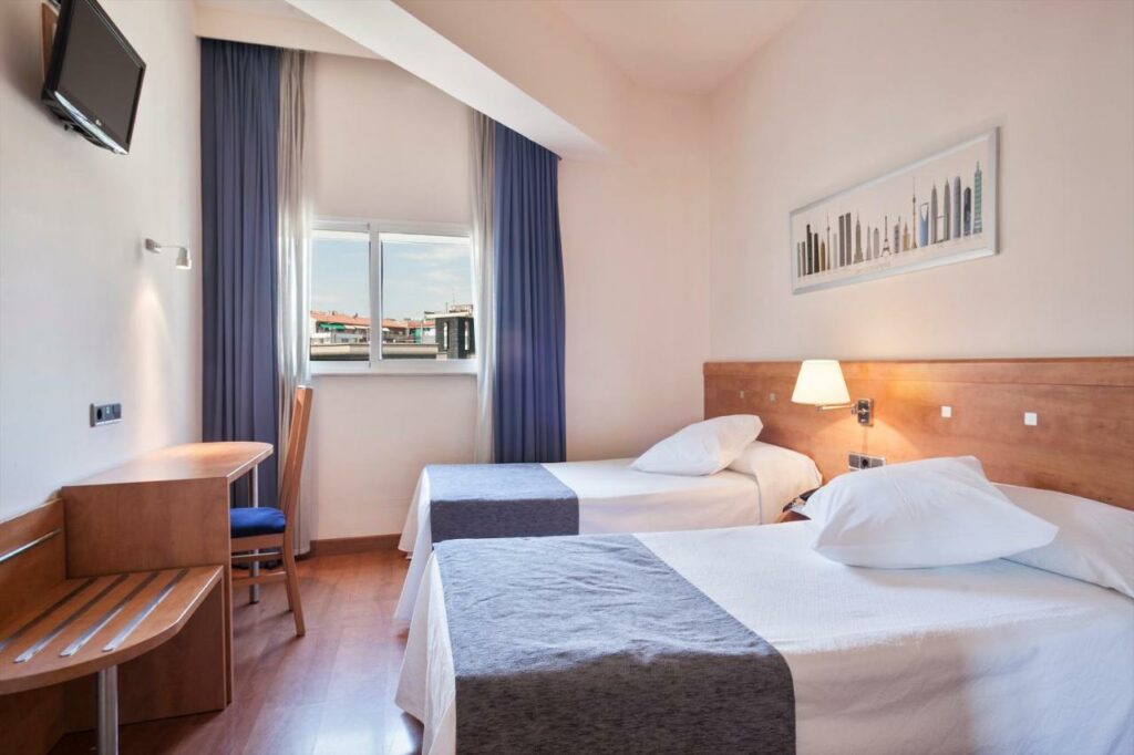 Quarto do Acta Antibes, uma das recomendações de hotéis perto da Sagrada Família em Barcelona. Duas camas de solteiro tem cabeceira de madeira atrás e uma mesinha com abajur no meio. De frente para elas há uma mesa com cadeira e uma televisão na parede acima. Ao fundo, uma janela tem cortinas azuis abertas. Há um quadro na parede acima das camas.