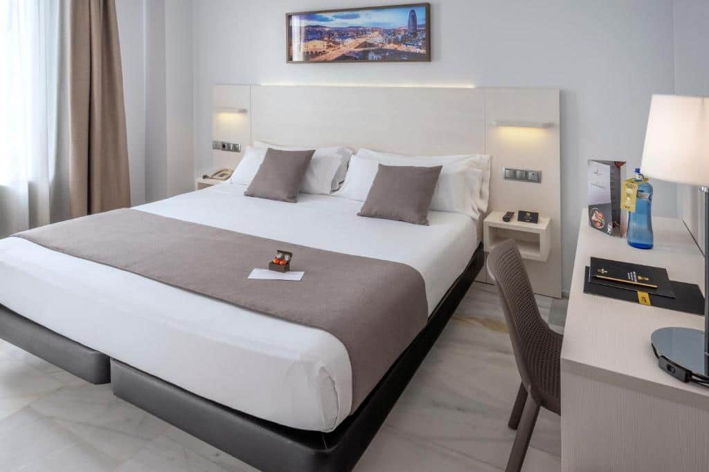 Quarto do Hotel Serhs Carlit, uma das recomendações de hotéis perto da Sagrada Família em Barcelona. A cama de casal tem mesinhas de cabeceira com luminárias dos dois lados. Do lado direito da cama, uma mesa de trabalho com cadeira, abajur e garrafa d'água está encostada na parede. Há um quadro na parede acima da cama.