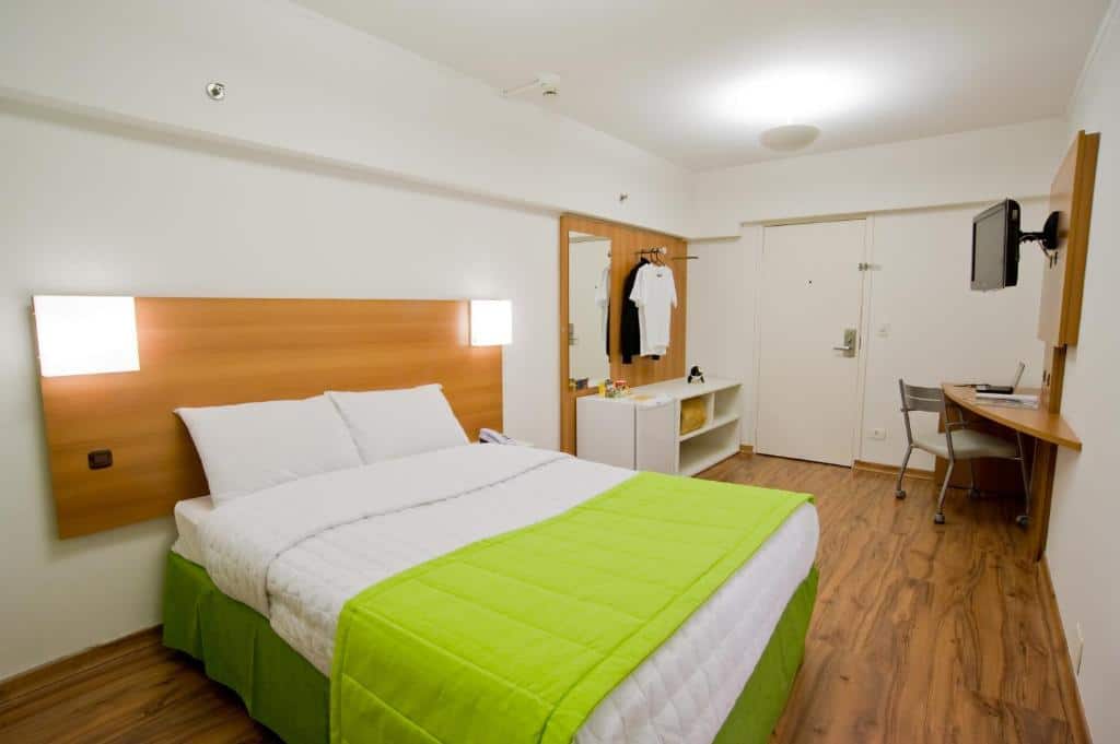 Quarto do Slim São Paulo Congonhas by Slaviero Hotéis com uma cama de casal com cobertas brancas e verde, chão de madeira, uma televisão, uma pequena mesa de canto com uma cadeira, um frigobar, um espelho e um cabide, para representar hotéis perto do Aeroporto de Congonhas