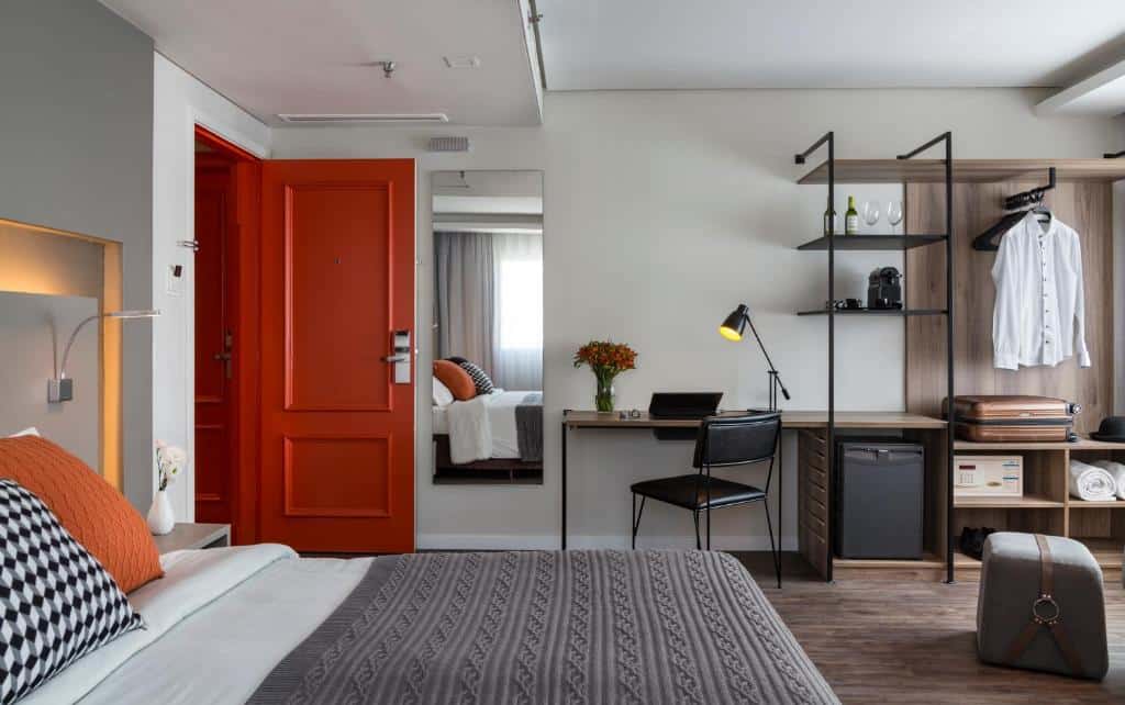 Quarto no Intercity São Paulo Ibirapuera com uma cama de casal, um espelho, uma mesa de escritório com uma cadeira e uma luminária, ao lado há uma estante com um frigobar e cafeteira, e um armário de conceito aberto, o chão é de madeira