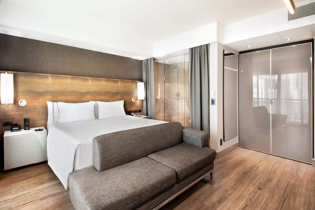 Quarto do Meliá Ibirapuera com uma cama de casal, do lado direito há um box de vidro com cortinas que leva o banheiro, ao lado há um armário de duas portas e um espelho, o chão é de madeira e na frente do sofá há um sofá, para representar os hotéis perto do Aeroporto de Congonhas