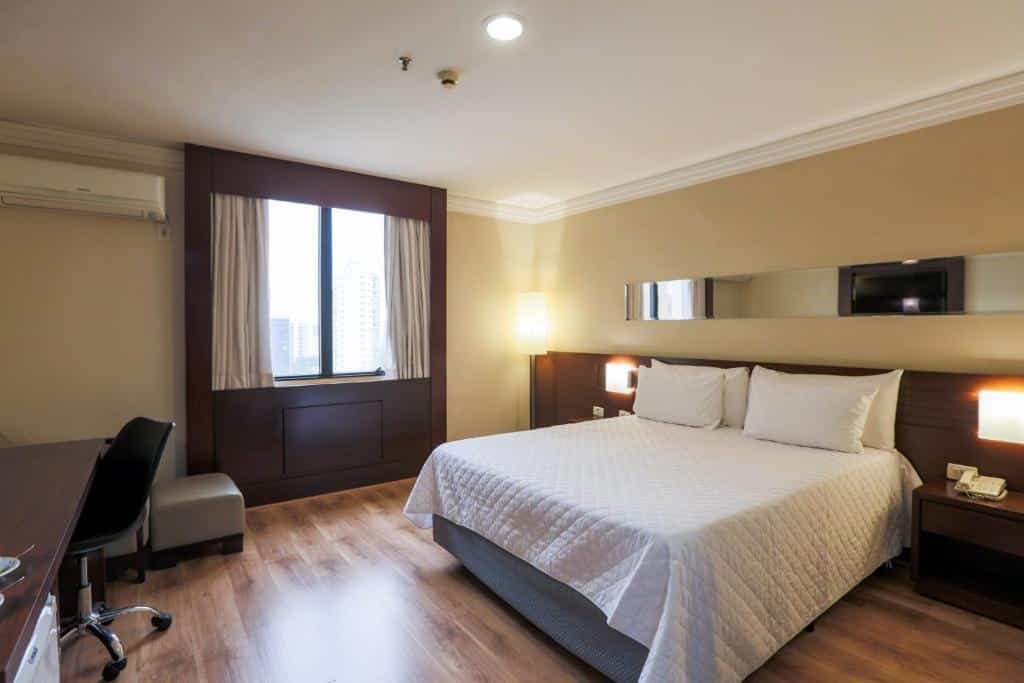 Quarto no Transamerica Executive Congonhas com uma cama de casal, uma janela com cortinas, uma cabeceira com iluminação indireta, uma mesa de escritório com cadeira giratória e um ar-condicionado