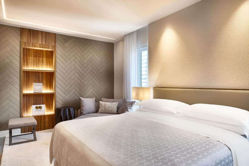 Quarto do Sheraton São Paulo WTC Hotel com uma cama de casal, uma poltrona ao lado da janela com cortinas, uma estante de madeira com iluminação indireta, um amplo tapete branco, para representar os hotéis perto do Aeroporto de Congonhas