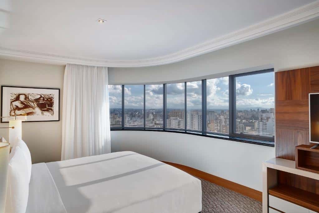 Quarto no Renaissance São Paulo Hotel com uma janela de vista panorâmica da cidade, uma cama de casal, chão de carpete, um raque com uma televisão