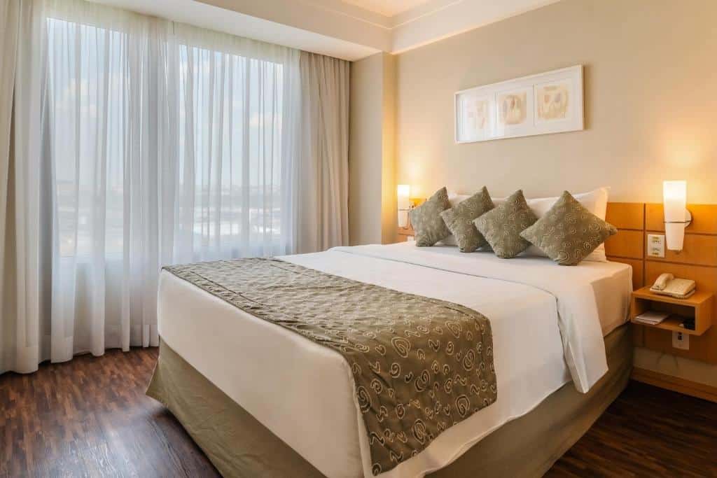 Uma cama do Mercure Guarulhos Aeroporto bem arrumada com quatro travesseiros, um tapete estampado, abajures de cabeceira, um telefone e uma grande janela com cortinas, para representar hotéis perto do Aeroporto de Guarulhos.