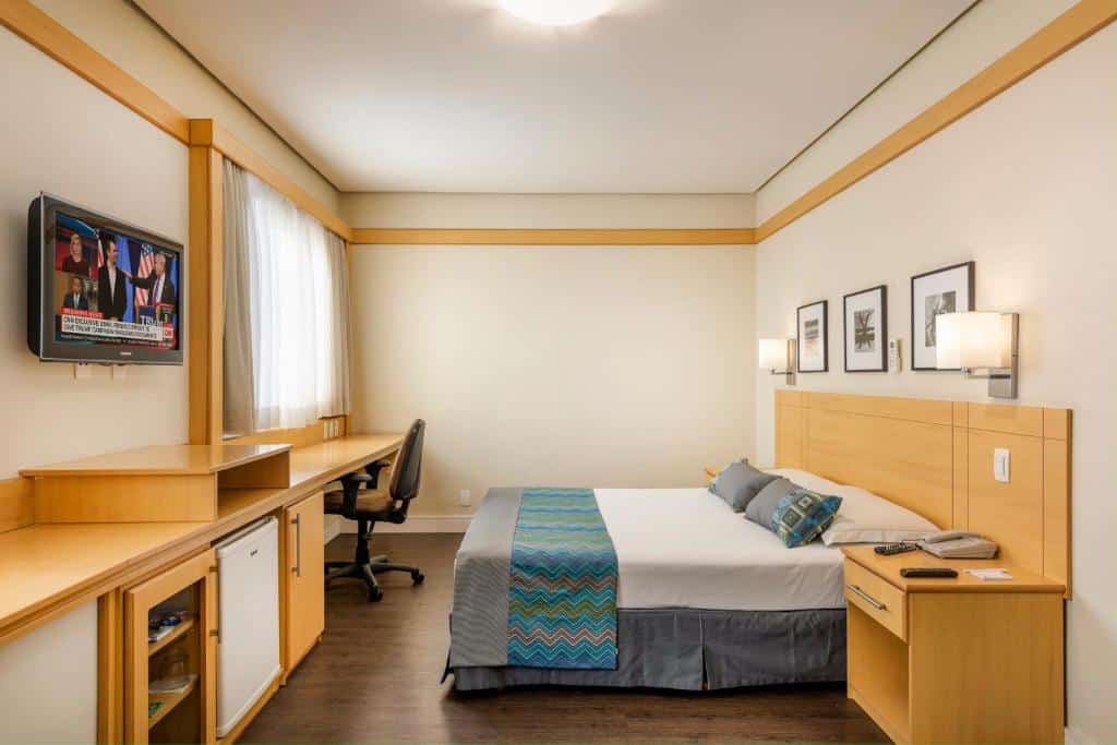 Quarto moderno no Monreale Plus Guarulhos International Airport com uma cama queen, mesa de trabalho, TV na parede, mesa de cabeceira e arte emoldurada em uma parede neutra, para representar hotéis perto do Aeroporto de Guarulhos.