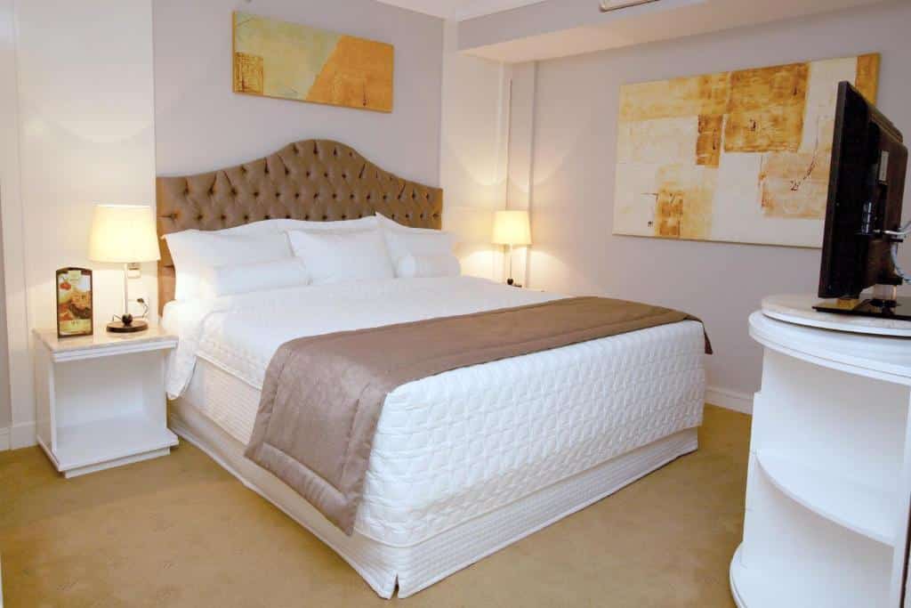 Quarto no Bristol International Airport Hotel com uma cama grande, duas mesinhas de cabeceira com luminárias, arte abstrata na parede e uma TV em um suporte branco, para representar hotéis perto do Aeroporto de Guarulhos.