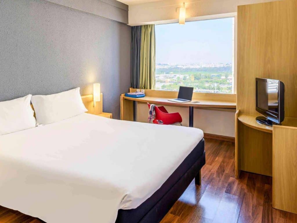 Quarto no ibis Guarulhos com uma cama de casal, TV pequena, escrivaninha com laptop, cadeira e uma janela grande com luz do dia do lado de fora, para rperesentar hotéis perto do Aeroporto de Guarulhos.
