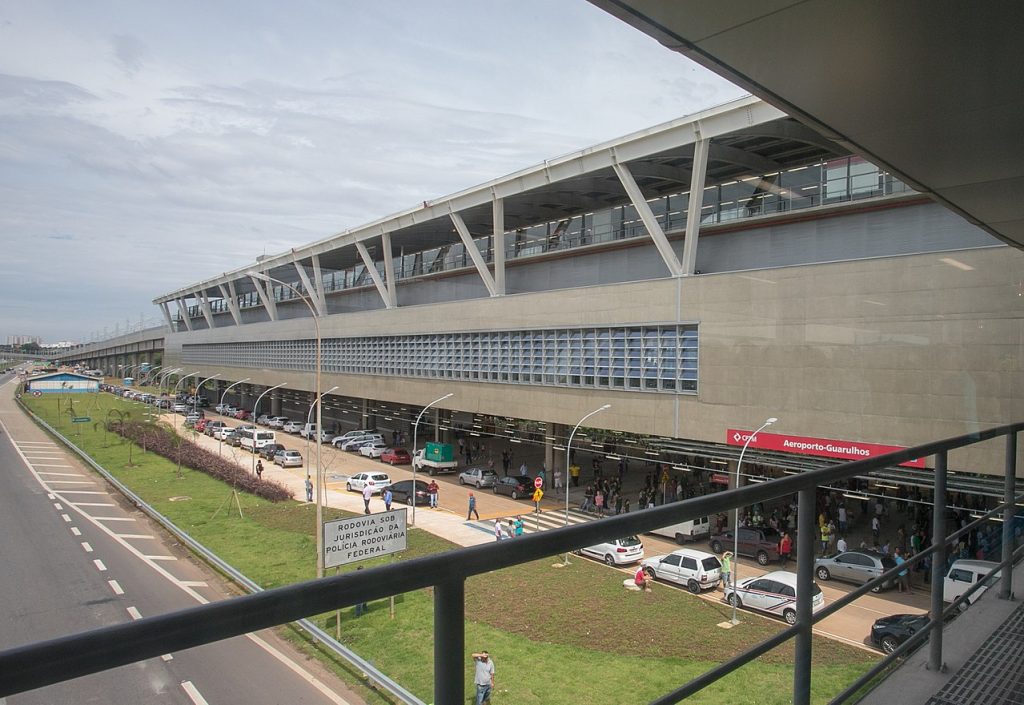 Vista externa de um terminal do Aeroporto de Guarulhos, movimentado com carros, pessoas e placas perto da entrada em um dia nublado.