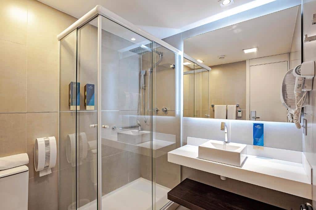 Banheiro moderno com box de vidro, pia branca, espelho grande, vaso sanitário e paredes de azulejos bege, no FAST SLEEP.