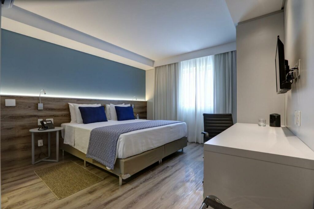 Quarto moderno no Comfort Hotel Guarulhos Aeroporto com uma cama de casal, detalhes em azul, escrivaninha, cadeira, TV na parede e janela grande com cortinas, para representar hotéis perto do Aeroporto de Guarulhos.