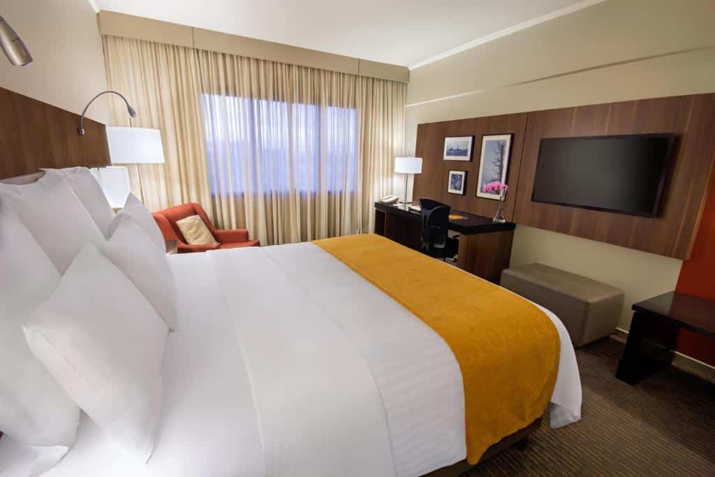 Quarto de casal em um dos hotéis perto do Aeroporto de Guarulhos, o Marriot São Paulo Airport Hotel, com TV plana grande, cama ampla, poltrona, abajur, mesa de trabalho e cortina.