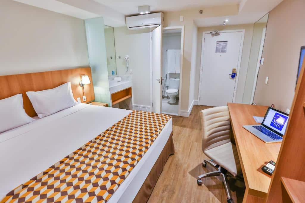 Quarto moderno no Sleep Inn Aeroporto de Guarulhos - São Paulo com uma cama de casal, mesa com laptop, cadeira e vista do banheiro e da porta de entrada, para representar hotéis perto do Aeroporto de Guarulhos.