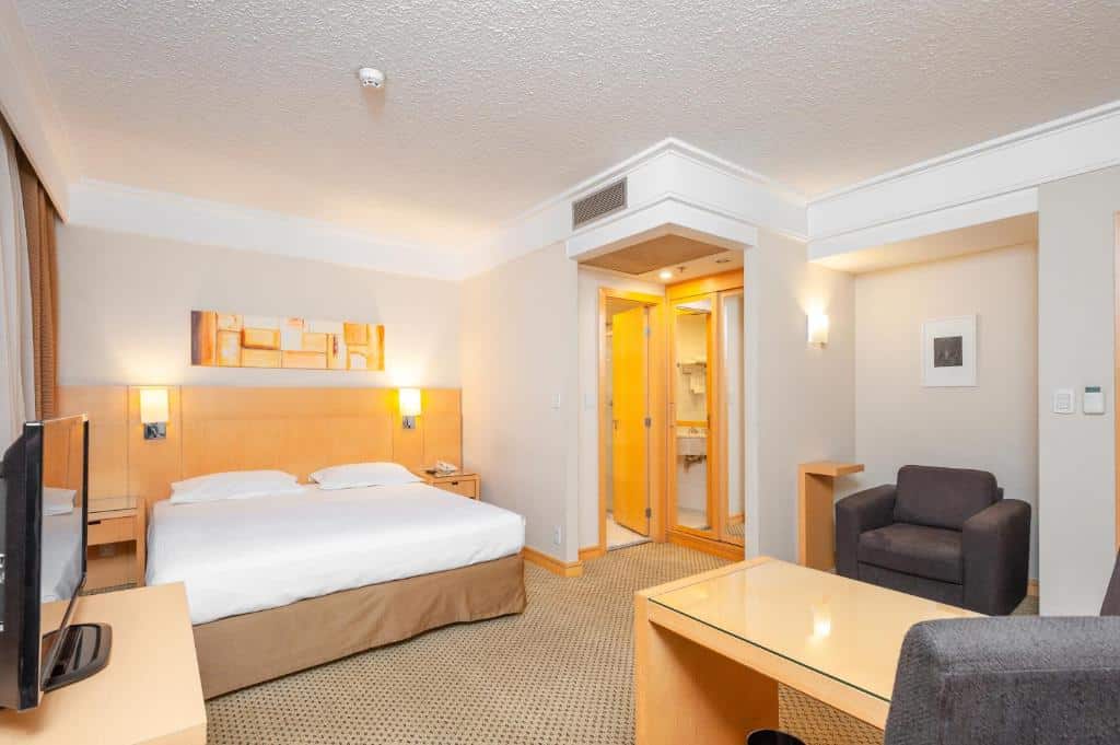 Um quarto no Slaviero Essential Guarulhos Aeroporto com uma cama de casal, uma TV, uma poltrona, uma escrivaninha e uma porta de banheiro aberta, para representar hotéis perto do Aeroporto de Guarulhos.