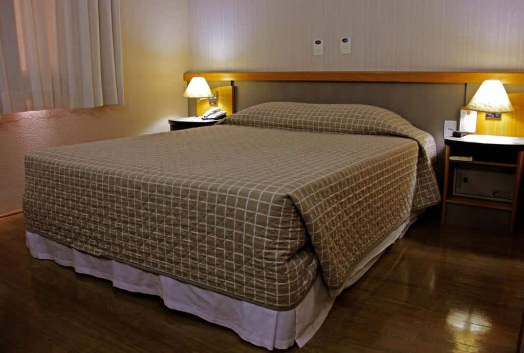 Um quartodo Sables Hotel Guarulhos com uma cama de casal bem arrumada, duas mesas de cabeceira, luminárias e um telefone em uma mesa, com piso de madeira, para representar hotéis perto do Aeroporto de Guarulhos.
