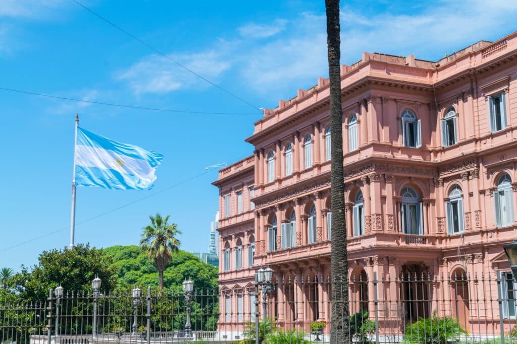 Prédio governamental rosa com janelas ornamentadas e um mastro com a bandeira argentina, em contraste com um céu azul e palmeiras, em Buenos Aires.