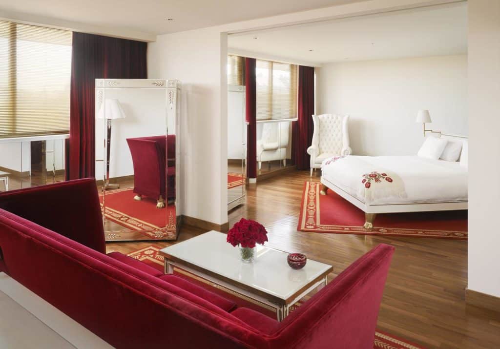 Quarto espaçoso no Faena Hotel Buenos Aires com uma cama branca, sofá de veludo vermelho, espelho grande e piso de madeira com tapetes vermelhos.