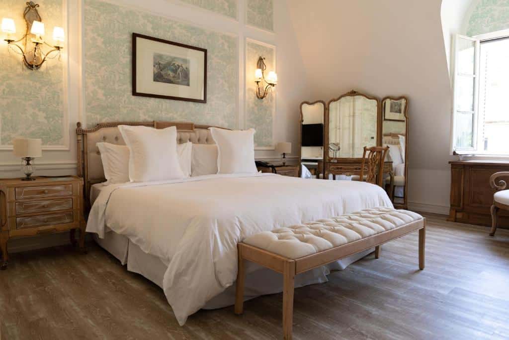 Uma cama bem arrumada com roupa de cama branca, móveis de madeira, um banco ao pé e uma janela aberta que deixa entrar a luz do dia, no Four Seasons Hotel Buenos Aires.