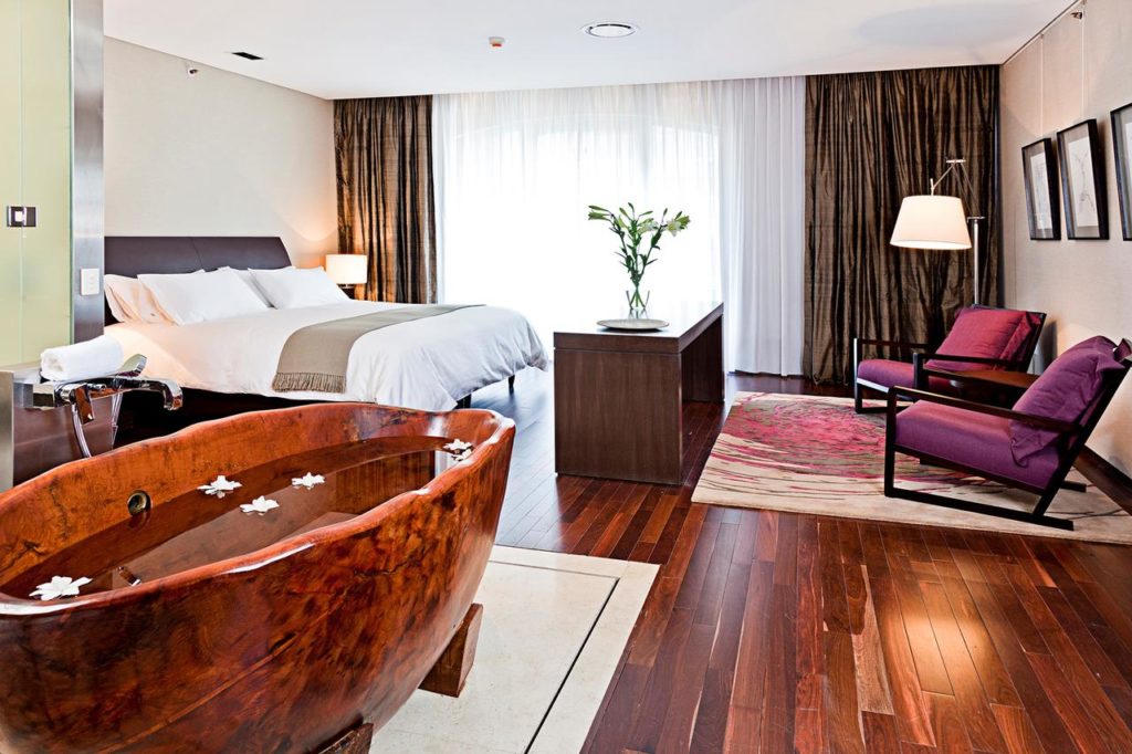 Quarto moderno no MIO Buenos Aires com banheira de madeira, cama de casal, duas cadeiras roxas e janela grande com cortinas transparentes.