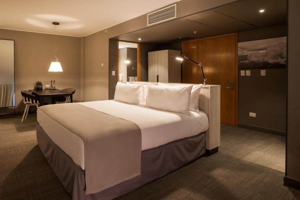 Quarto do Solace Hotel Santiago moderno com uma cama grande, luminárias de cabeceira, uma mesa com cadeiras e decoração em cores neutras.
