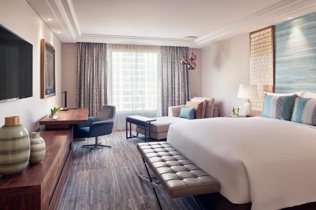 Quarto do The Ritz-Carlton com cama de casal do lado direito em frente a cama, TV e do lado esquerdo do quarto poltrona e mesa de trabalho.