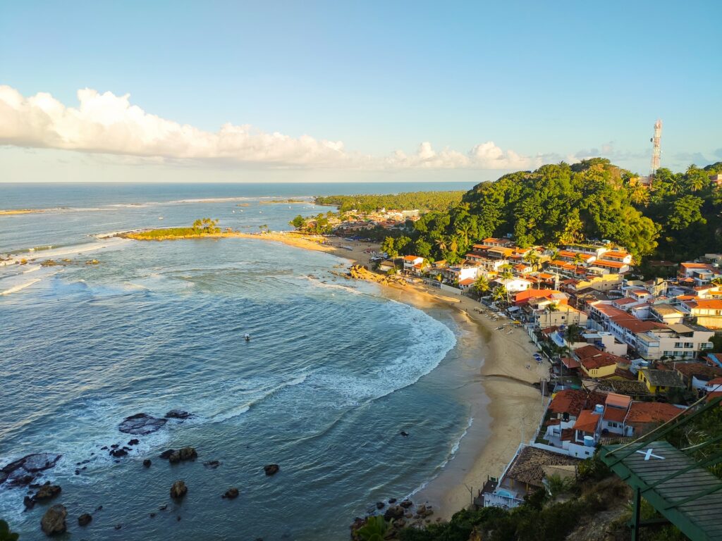 Vista aérea de uma cidade costeira com praias arenosas, litoral rochoso, oceano azul e casas cercadas por colinas verdejantes.