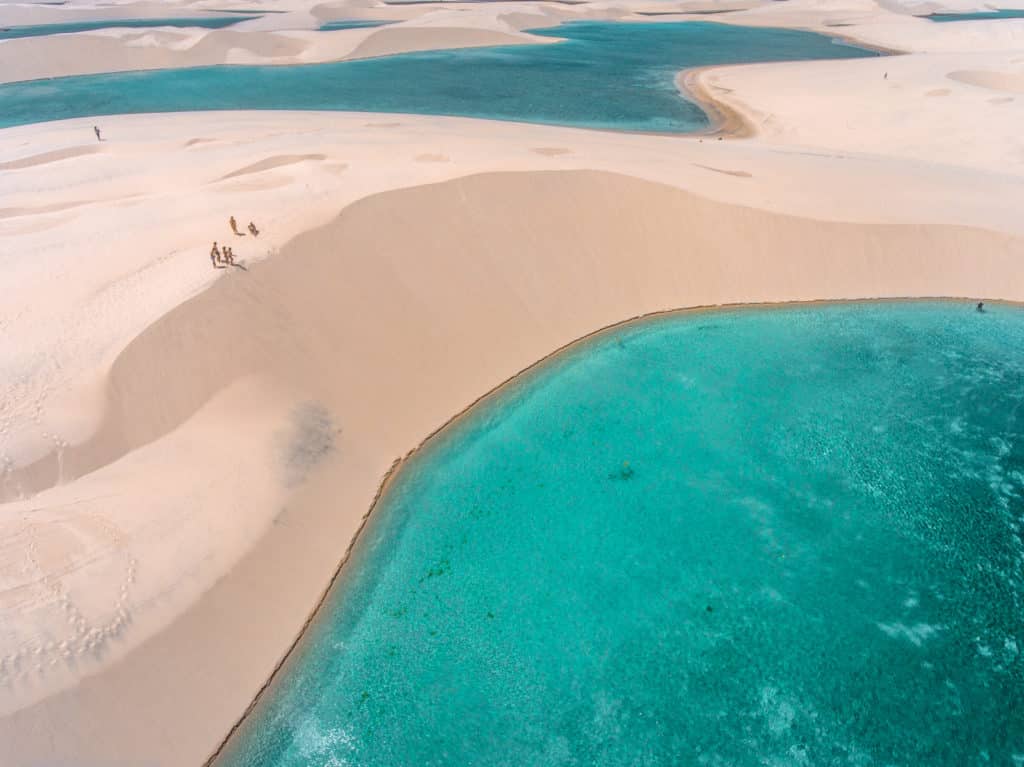 Vista aérea de dunas de areia e lagoas azul-turquesa no Parque Nacional dos Lençóis Maranhenses, no Brasil, com algumas pessoas caminhando.