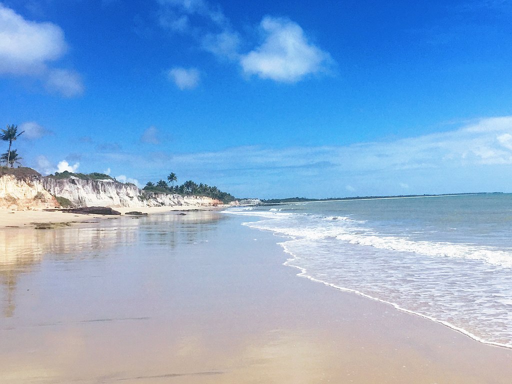Praia de areia com ondas suaves, céu azul e uma fileira de penhascos e árvores ao longe, sob nuvens brancas dispersas.