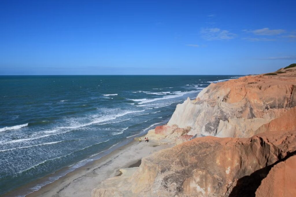 Vista do penhasco de uma praia com ondas tocando a costa sob um céu azul claro. Duas pessoas estão caminhando perto da água.