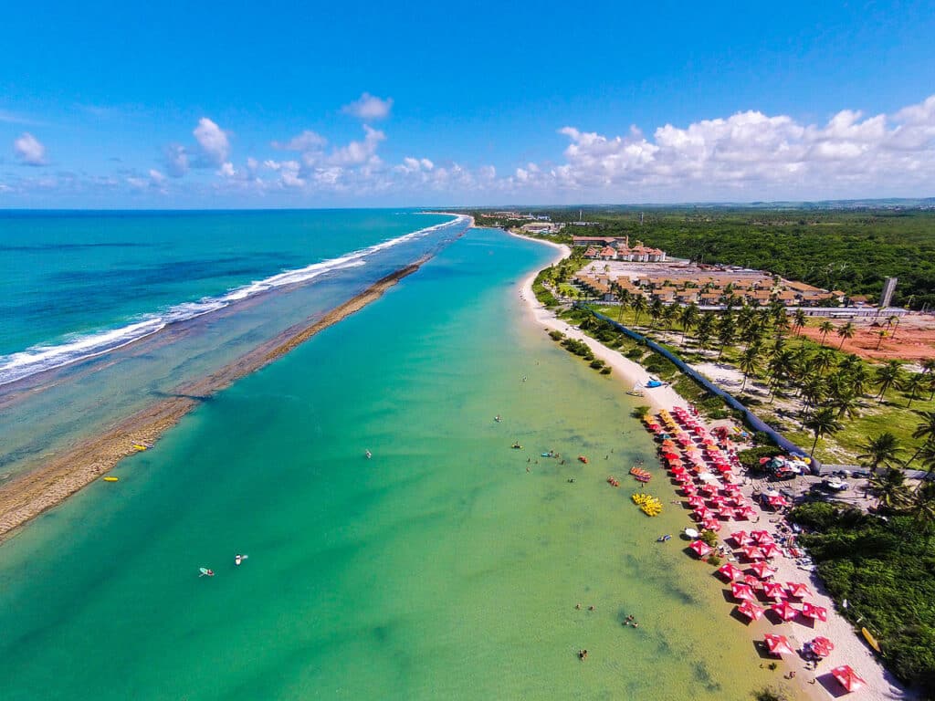 Vista aérea de uma praia tropical com águas cristalinas azul-turquesa, uma praia arenosa repleta de guarda-sóis e um complexo hoteleiro distante.