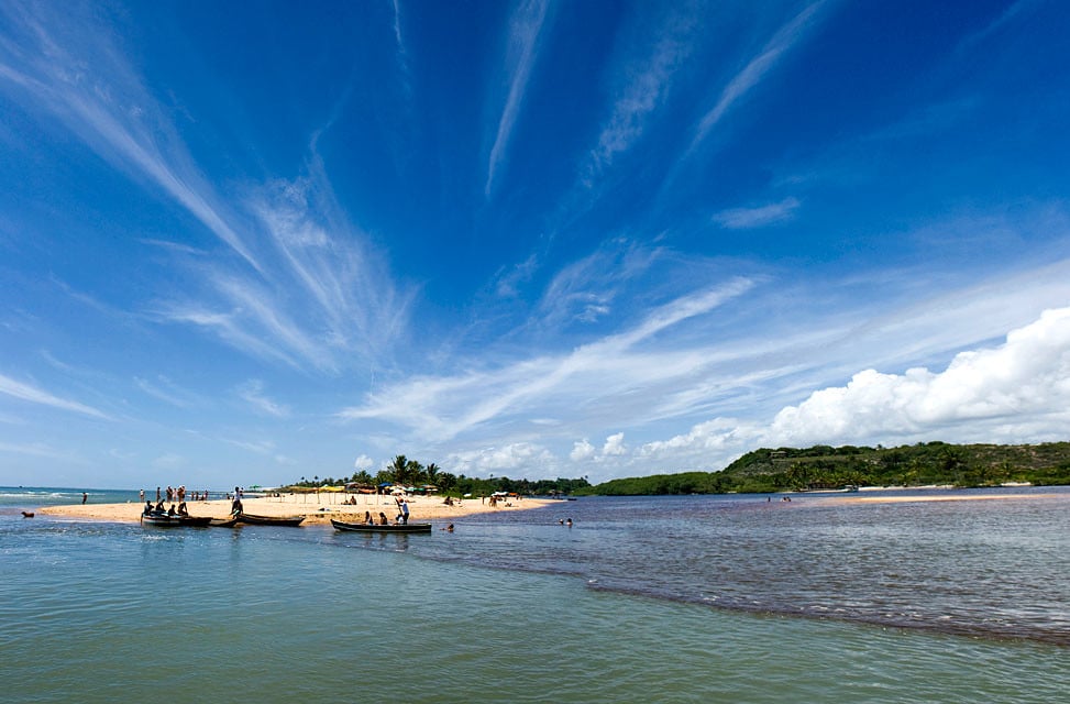 Pequenos barcos atracados em uma ilha arenosa com palmeiras, cercada por água sob um céu azul com nuvens finas.