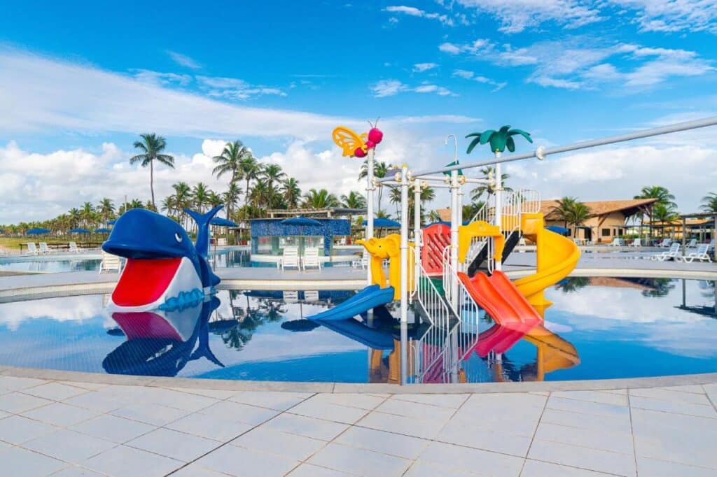Uma área de recreação aquática para crianças com toboáguas coloridos e um brinquedo de baleia, situada em uma piscina com palmeiras e prédios do Makai Resort All Inclusive Convention Aracaju ao fundo.
