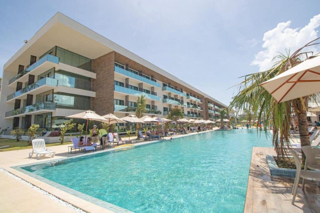 Grande piscina ao ar livre no Maceio Mar Resort All Inclusive com guarda-sóis e espreguiçadeiras ao lado de um moderno edifício de quatro andares sob um céu parcialmente nublado.