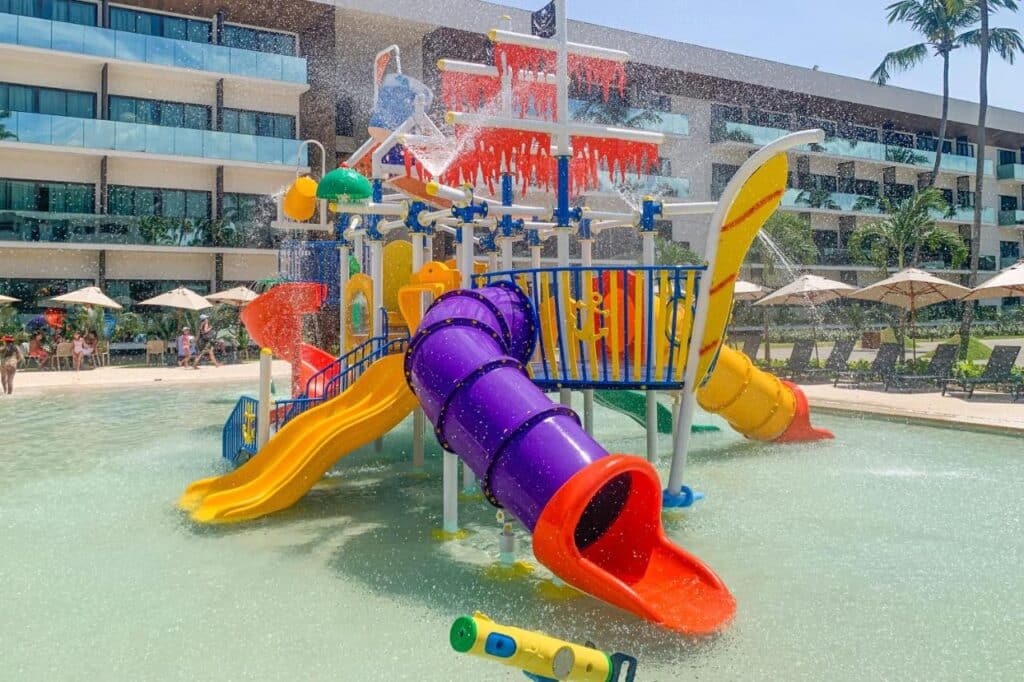 Parque aquático colorido no Maceio Mar Resort All Inclusive com toboáguas, jatos d'água e brinquedos aquáticos em uma piscina rasa, cercada por espreguiçadeiras e guarda-sóis.