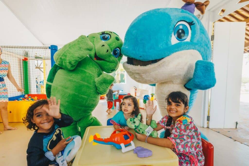 Na imagem, três crianças estão sentadas ao redor de uma mesa de brinquedos, sorrindo e acenando para a câmera no Sauipe Resorts Ala Terra - All Inclusive, um dos melhores resorts all inclusive no Brasil. Elas estão acompanhadas por duas mascotes fantasiadas: uma tartaruga verde e uma baleia azul, que estão interagindo de forma amigável com as crianças. O ambiente é claro e colorido, com uma área de recreação ao fundo, onde há uma piscina de bolinhas.