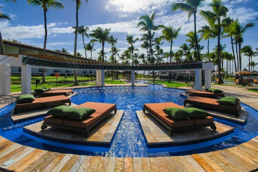 A imagem mostra uma área de piscina do Transamerica Comandatuba All Inclusive Resort, um dos melhores resorts all inclusive no Brasil. No centro da piscina, há várias espreguiçadeiras com almofadas verdes, posicionadas em plataformas de pedra dentro da água. Ao fundo, há uma estrutura de madeira e metal com cascatas de água que caem na piscina. A área ao redor é cercada por palmeiras altas e um céu azul com poucas nuvens, criando um ambiente tropical e relaxante. Ao redor da piscina, existem áreas verdes bem cuidadas e outras espreguiçadeiras dispostas para os hóspedes aproveitarem o sol.