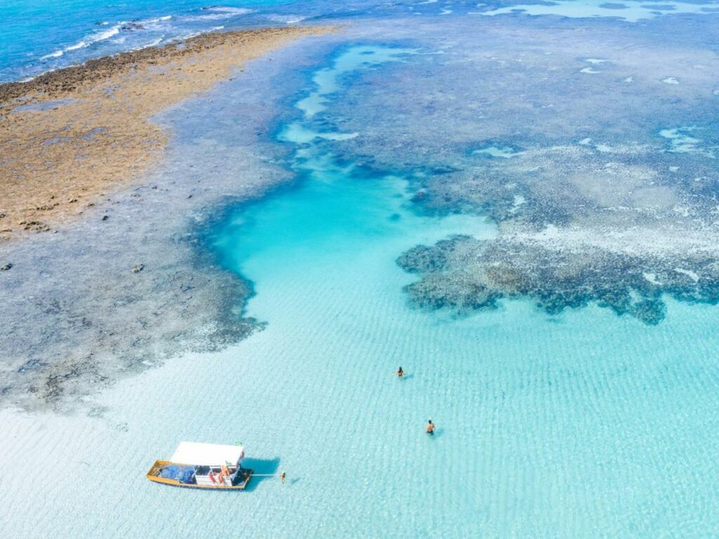 A imagem mostra uma bela vista aérea de um trecho de recifes de corais em uma área de águas rasas e cristalinas, próximo ao Japaratinga Lounge Resort. No centro da imagem, há um pequeno barco com uma cobertura branca flutuando sobre a água turquesa, cercado por duas pessoas nadando. As cores variam entre tons de azul e verde, com o recife de corais criando um contraste interessante com as águas claras. A paisagem natural é serena e convida ao relaxamento e exploração subaquática, destacando a beleza exuberante da região costeira.