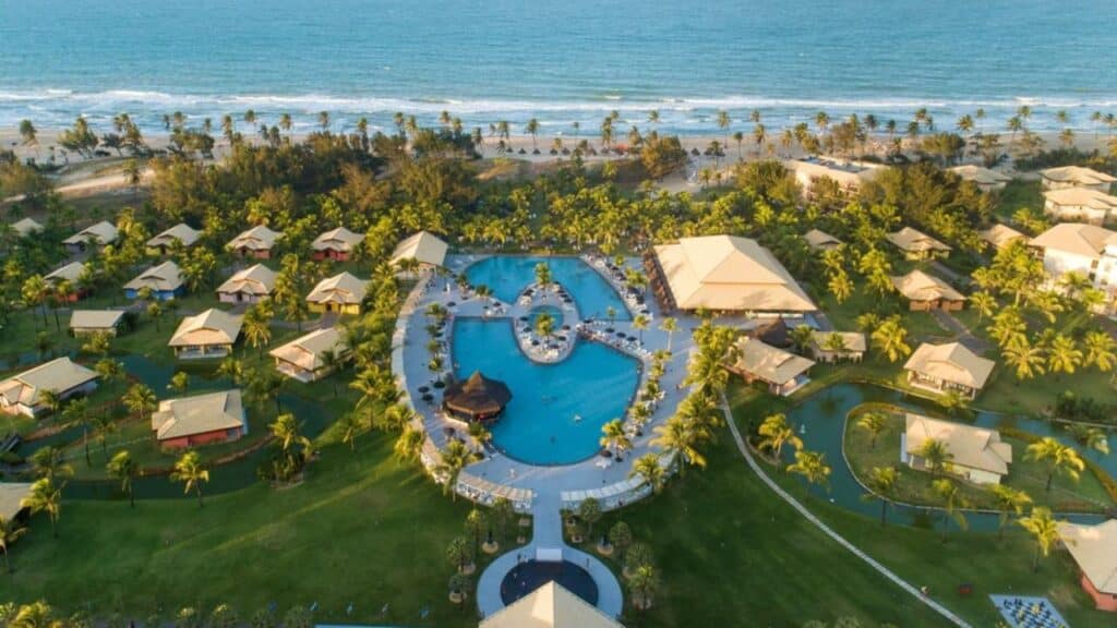 A imagem mostra uma vista aérea do Vila Galé Resort Cumbuco, com uma ampla área de lazer e acomodação. No centro, há uma grande piscina em formato irregular, cercada por espreguiçadeiras e guarda-sóis. Ao redor da piscina, existem várias construções com telhados inclinados, que parecem ser bangalôs ou chalés. Mais ao fundo, é possível ver a praia com sua areia branca e o mar azul, cercado por coqueiros que acrescentam um charme tropical ao cenário. O resort está rodeado por vegetação exuberante, criando um ambiente paradisíaco e tranquilo para os hóspedes.