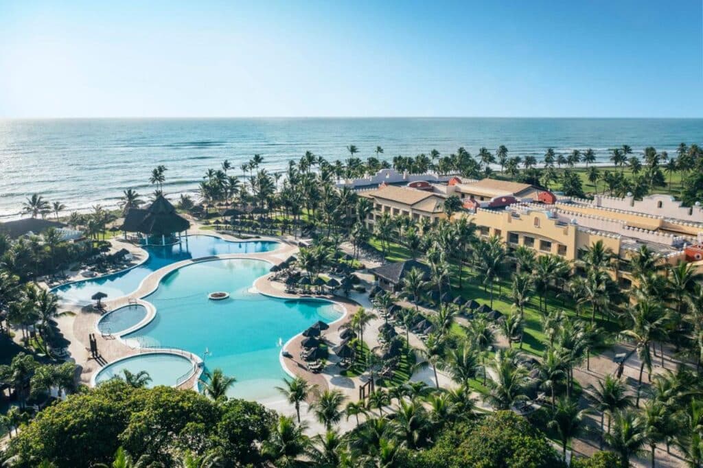 Vista aérea do Iberostar Waves Bahia - All Inclusive à beira-mar com uma piscina em formato de lagoa, cercado por palmeiras e edifícios, perto do oceano.