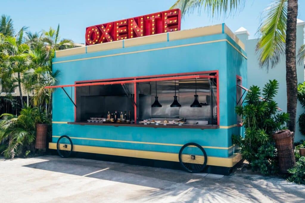 Uma barraca de comida azul e amarela com uma placa vermelha "OXENTE" no topo, cercada por palmeiras e plantas no Iberostar Waves Bahia - All Inclusive.